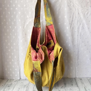 May include: Mustard yellow tote bag with patterned straps and a pink paisley interior. The bag is hanging from a black hook against a white background. The straps feature a mix of yellow, blue, and red patterns.