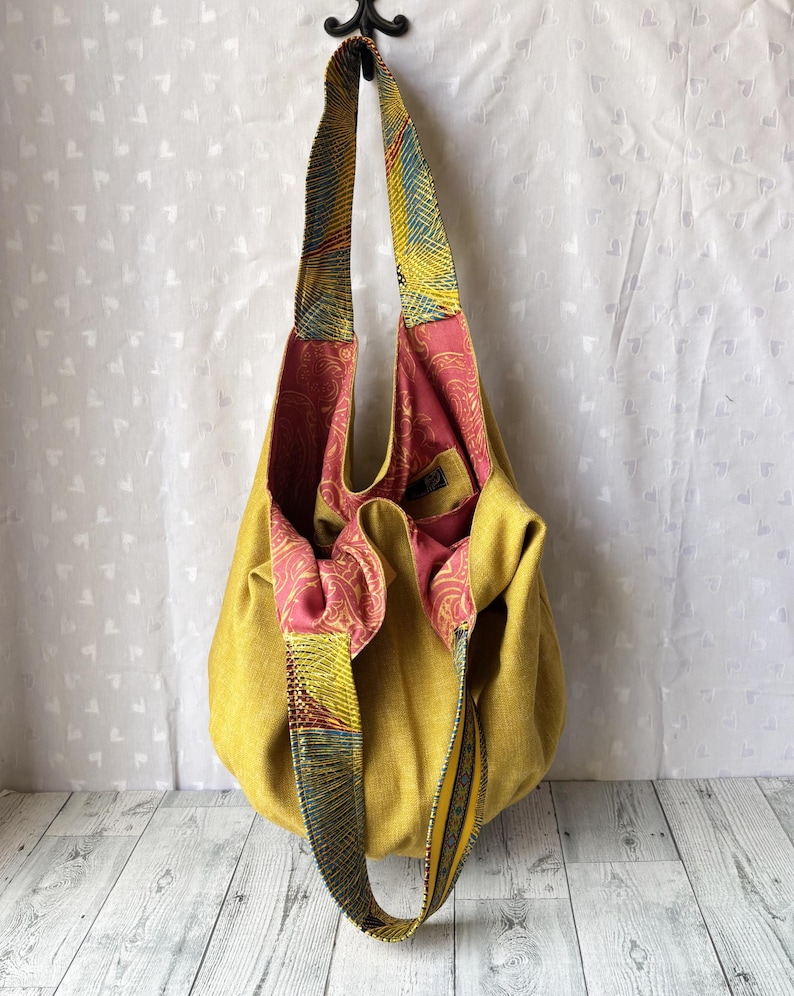 May include: Mustard yellow tote bag with patterned straps and a pink paisley interior. The bag is hanging from a black hook against a white background. The straps feature a mix of yellow, blue, and red patterns.