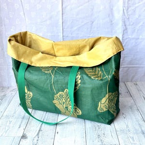 May include: A rectangular tote bag featuring a dark green exterior with a gold floral pattern and two green fabric handles. The interior lining is a solid mustard yellow fabric that folds over the top edge of the bag.