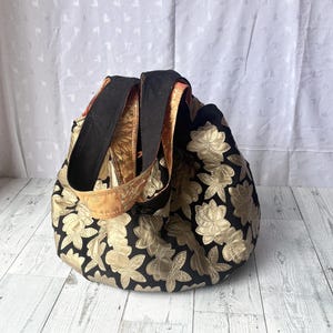 Op de afbeelding: Een zwarte tote bag met gouden bloemenborduurwerk. De tas heeft twee lange hengsels.