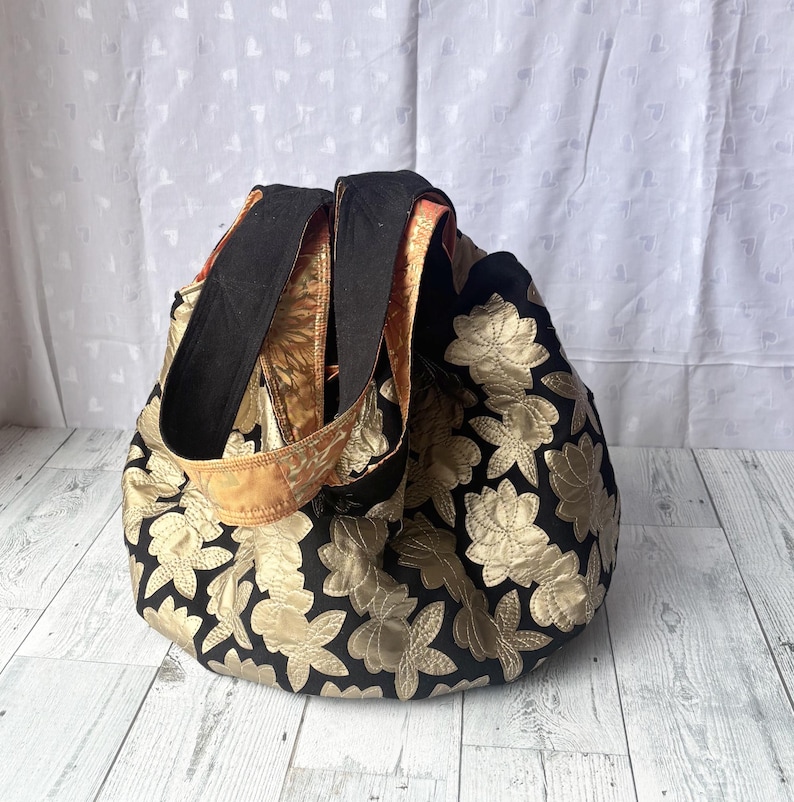 Op de afbeelding: Een zwarte tote bag met gouden bloemenborduurwerk. De tas heeft twee lange hengsels.