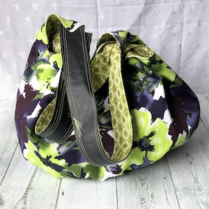 May include: A large, green and purple floral print tote bag with black leather straps. The bag is made of fabric and has a casual, relaxed style.