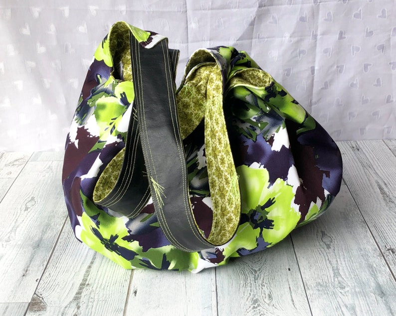 May include: A large, green and purple floral print tote bag with black leather straps. The bag is made of fabric and has a casual, relaxed style.