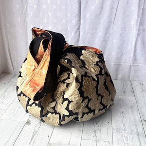 Op de afbeelding: Een zwarte tote bag met een gouden bloemenpatroon. De tas heeft een lange band en is gemaakt van een zachte, gewatteerde stof.