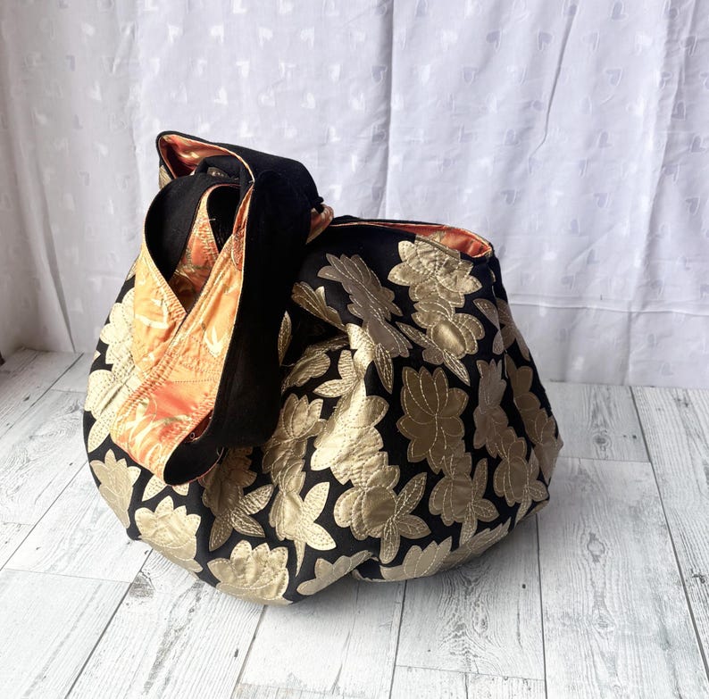 Op de afbeelding: Een zwarte tote bag met een gouden bloemenpatroon. De tas heeft een lange band en is gemaakt van een zachte, gewatteerde stof.