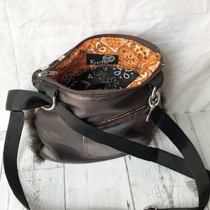 Pu&ograve; includere: Una borsa a tracolla in pelle marrone con una tracolla nera e una fodera in bandana nero e arancione. La borsa ha una piccola tasca sul davanti e una zip argentata. Il nome del marchio "KuChI" &egrave; visibile sulla fodera.