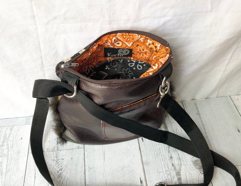 Pu&ograve; includere: Una borsa a tracolla in pelle marrone con una tracolla nera e una fodera in bandana nero e arancione. La borsa ha una piccola tasca sul davanti e una zip argentata. Il nome del marchio "KuChI" &egrave; visibile sulla fodera.
