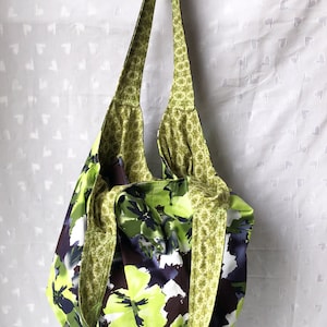 May include: A large green and white floral print tote bag with a green and gold patterned strap. The bag is hanging from a hook.