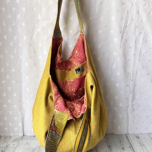 May include: A mustard yellow shoulder bag with a patterned strap and interior. The bag's interior features a pink and red floral design. The strap has a colorful geometric pattern. The bag is made of a soft, textured fabric.