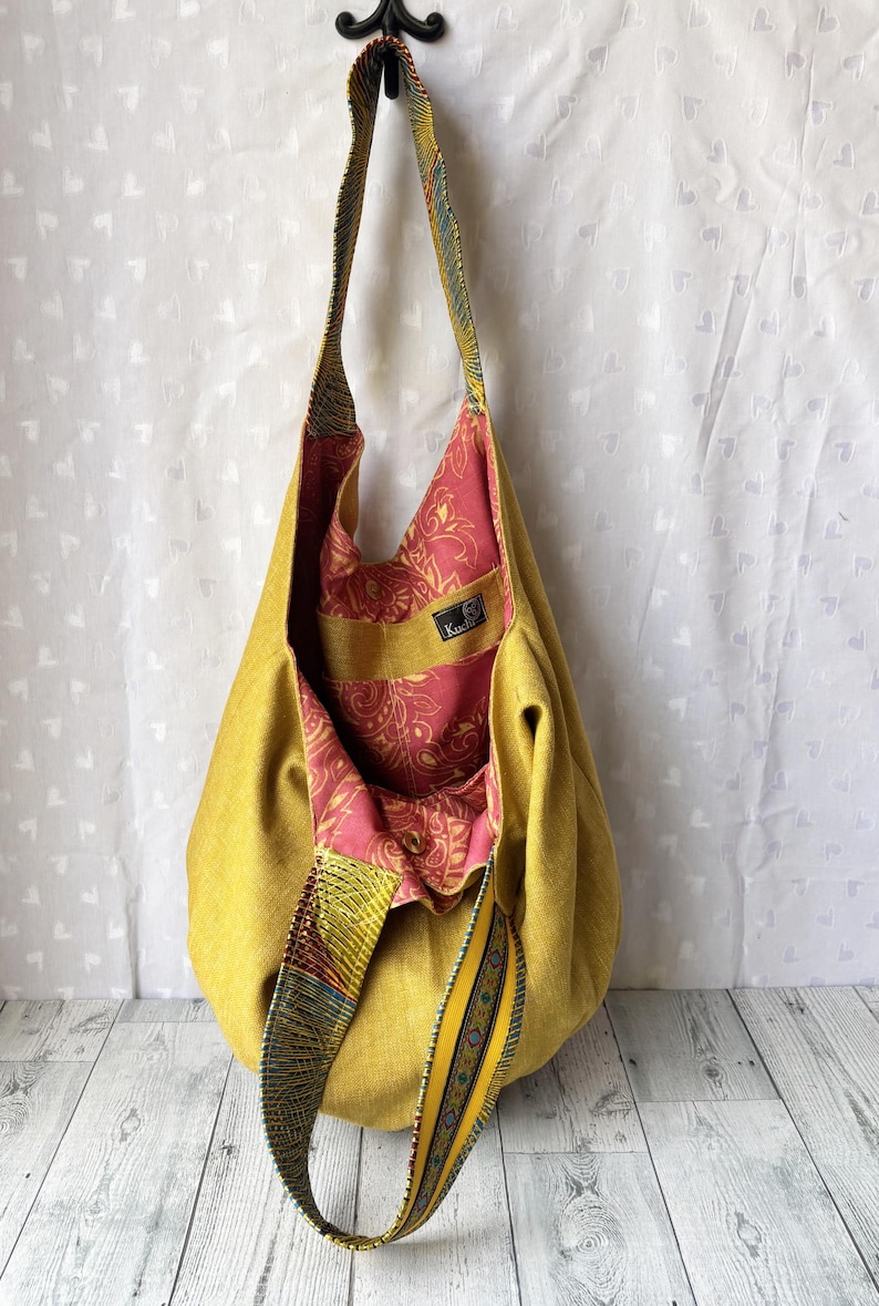 May include: A mustard yellow shoulder bag with a patterned strap and interior. The bag's interior features a pink and red floral design. The strap has a colorful geometric pattern. The bag is made of a soft, textured fabric.