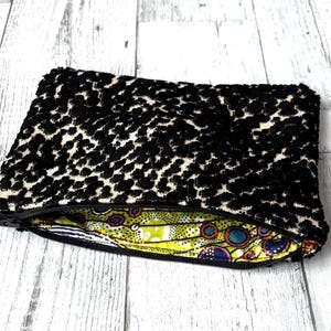 May include: A black and cream-colored leopard print clutch bag with a zipper closure. The interior lining features a colorful, patterned fabric with yellow, blue, and red accents. The bag is rectangular and appears to be made of a soft, textured material.