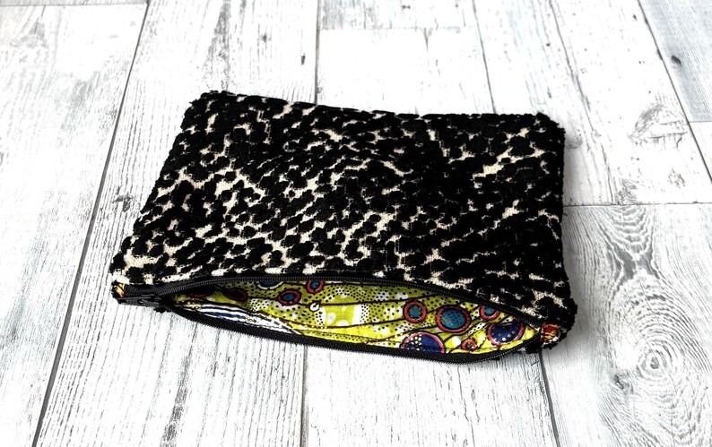 May include: A black and cream-colored leopard print clutch bag with a zipper closure. The interior lining features a colorful, patterned fabric with yellow, blue, and red accents. The bag is rectangular and appears to be made of a soft, textured material.
