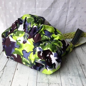 May include: A green, purple, and brown floral print tote bag with black straps. The bag is made of fabric and has a gathered top.