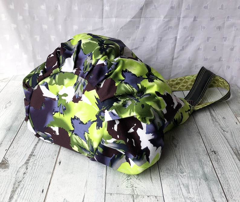May include: A green, purple, and brown floral print tote bag with black straps. The bag is made of fabric and has a gathered top.