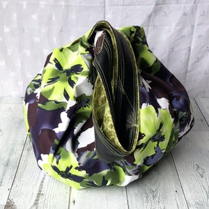 May include: A large, green and white floral print tote bag with a black and green strap. The bag is made of fabric and has a casual, bohemian style.