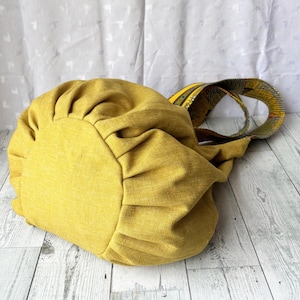 May include: Mustard yellow tote bag with a gathered design. The bag has a patterned strap in shades of yellow, green, and black. The bag is made of a textured fabric and sits on a light wood surface.