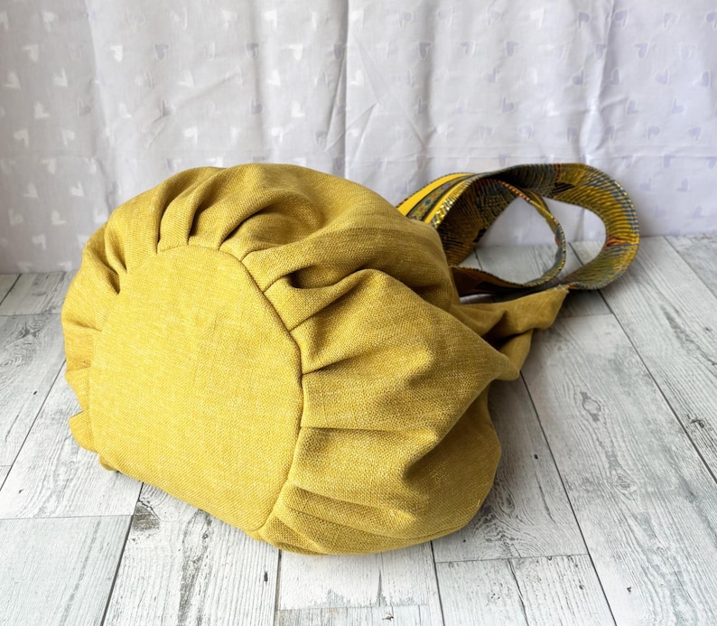 May include: Mustard yellow tote bag with a gathered design. The bag has a patterned strap in shades of yellow, green, and black. The bag is made of a textured fabric and sits on a light wood surface.