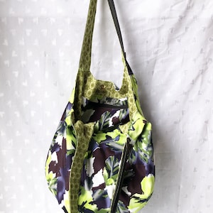 May include: A large green and purple floral print tote bag with a black zipper and a black strap. The bag is hanging from a hook.