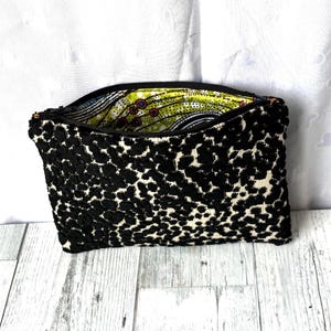 May include: Black and white patterned clutch bag with a zipper closure. The exterior features a textured, spotted design, while the interior lining displays a colorful, patterned fabric. The bag is open, revealing the vibrant interior.