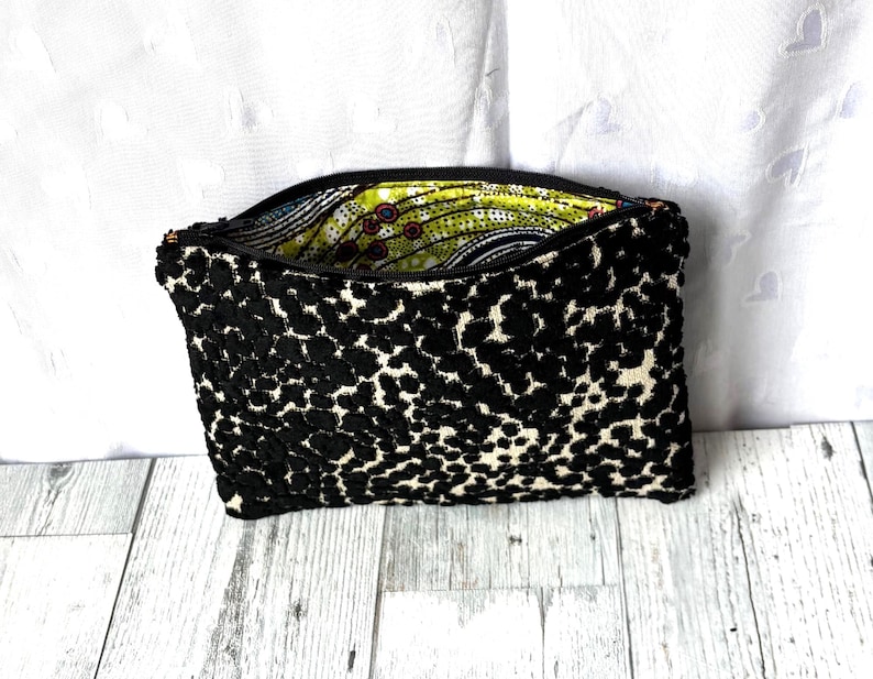 May include: Black and white patterned clutch bag with a zipper closure. The exterior features a textured, spotted design, while the interior lining displays a colorful, patterned fabric. The bag is open, revealing the vibrant interior.