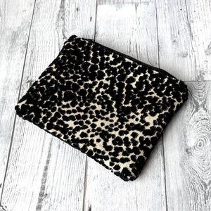 May include: A rectangular black and beige leopard print clutch with a black zipper. The clutch is made of a textured fabric and is set against a light gray wooden background. The bag is closed.