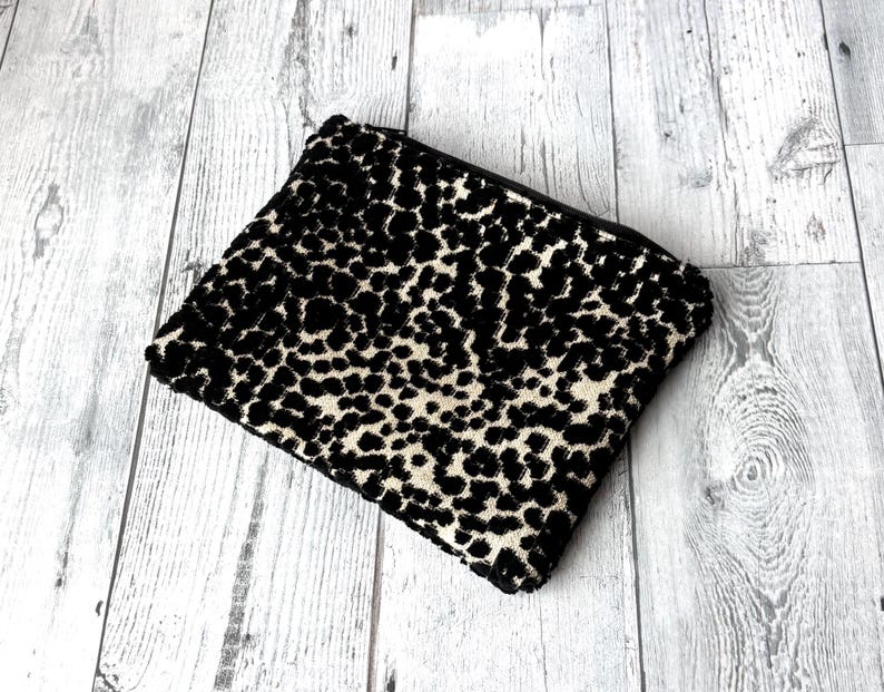 May include: A rectangular black and beige leopard print clutch with a black zipper. The clutch is made of a textured fabric and is set against a light gray wooden background. The bag is closed.