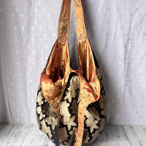 Op de afbeelding: Een grote zwarte tote bag met een gouden bloemenpatroon. De tas heeft een lange band en is gemaakt van een zachte, gestructureerde stof. De binnenkant van de tas is gevoerd met een feloranje stof.