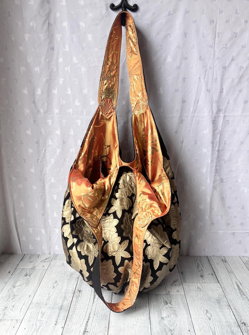 Op de afbeelding: Een grote zwarte tote bag met een gouden bloemenpatroon. De tas heeft een lange band en is gemaakt van een zachte, gestructureerde stof. De binnenkant van de tas is gevoerd met een feloranje stof.