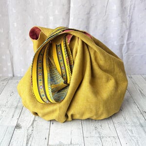 May include: Mustard yellow fabric bag with a decorative strap. The strap features a yellow background with blue and green geometric patterns. The bag's interior is a deep red color. The bag is sitting on a light wood surface.