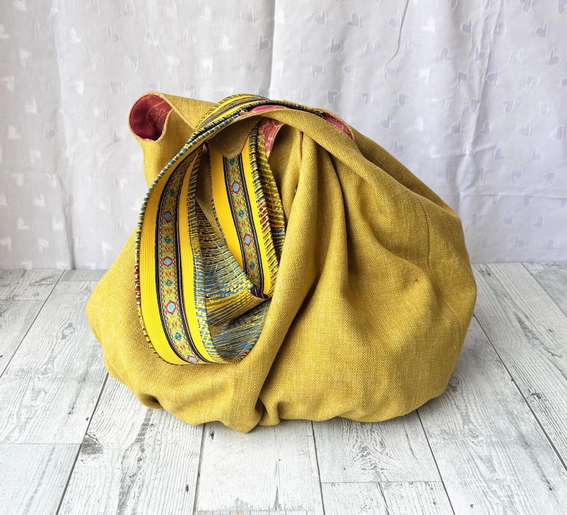 May include: Mustard yellow fabric bag with a decorative strap. The strap features a yellow background with blue and green geometric patterns. The bag's interior is a deep red color. The bag is sitting on a light wood surface.