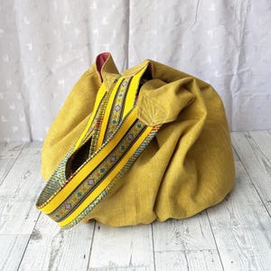 May include: Mustard yellow fabric tote bag with a decorative strap. The strap is yellow with a patterned design in blue, red, and green. The bag has a rounded shape and a contrasting pink interior.