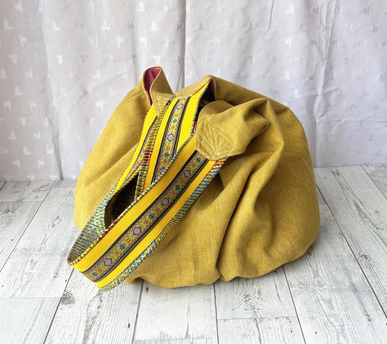 May include: Mustard yellow fabric tote bag with a decorative strap. The strap is yellow with a patterned design in blue, red, and green. The bag has a rounded shape and a contrasting pink interior.