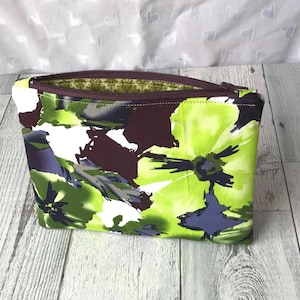 May include: A rectangular fabric zipper pouch featuring a bold, abstract floral print in shades of lime green, deep purple, and white. The pouch has a dark purple zipper closure and an interior lining with a green and white pattern.