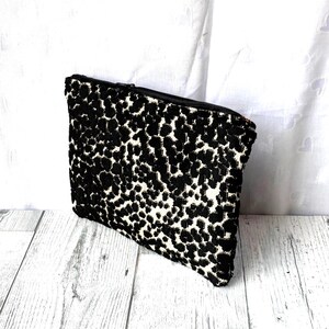 May include: A rectangular black and white patterned clutch bag with a zipper closure. The bag features a repeating pattern of black spots on a white background. The zipper is black, and the bag is made of a textured fabric.