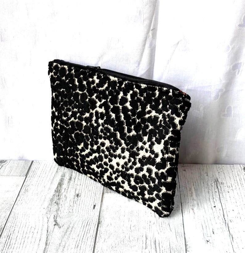 May include: A rectangular black and white patterned clutch bag with a zipper closure. The bag features a repeating pattern of black spots on a white background. The zipper is black, and the bag is made of a textured fabric.