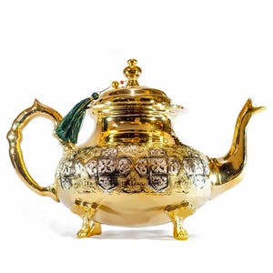 May include: A polished gold-tone teapot featuring an ornate embossed pattern with silver-colored accents on the body. The teapot has a curved handle, a long spout, three decorative feet, a lid with a finial, and a small dark green tassel.