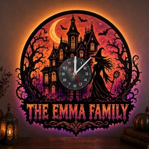 Op de afbeelding: Een ronde wandklok met een verlicht silhouet van een spookhuis, een maansikkel, vliegende vleermuizen en een heks met een staf. De tekst THE EMMA FAMILY staat onderaan gedrukt. De klok heeft witte wijzers op de wijzerplaat.