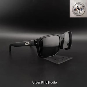 Könnte beinhalten: Schwarze Sonnenbrille mit rechteckigem Rahmen und dunkel getönten Gläsern. Die Bügel zeigen ein silberfarbenes, rechteckiges Logo-Emblem in der Nähe des Scharniers, und zwei kleine silberfarbene Nieten sind an den vorderen Rahmenecken sichtbar.