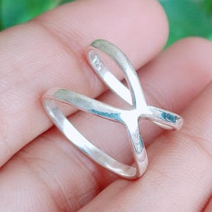 Peut inclure: Une bague en argent poli présentant un design croisé formant une forme en X. L'anneau métallique possède une surface lisse et réfléchissante et comporte un petit poinçon 925 sur le côté intérieur, indiquant la composition en argent sterling.