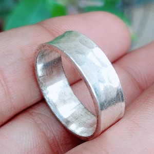 May include: A silver-colored ring with a wide band and a hammered, textured finish. The metal surface reflects light across its uneven, dimpled exterior, creating a metallic sheen. The band has a consistent width and a rounded, circular shape.