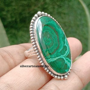 May include: An oval malachite gemstone ring set in a sterling silver bezel with a beaded edge. The green stone features natural concentric banding patterns. The text silverartsianstudio is visible on the surface of the ring setting.