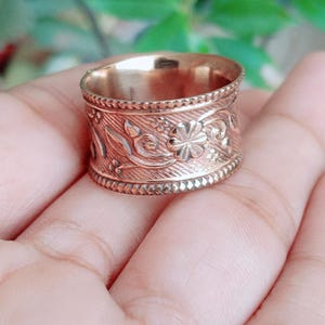 Può includere: Un anello largo in metallo color rame caratterizzato da un intricato motivo floreale in rilievo sulla fascia centrale. I bordi superiore e inferiore dell'anello sono rifiniti con una delicata texture a perline, creando un design uniforme lungo tutta la circonferenza.