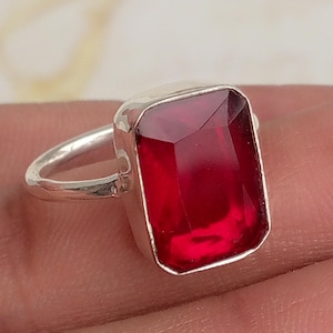 May include: A silver ring featuring a large, rectangular, faceted red gemstone set in a simple metal band. The stone has a deep crimson hue and a polished surface, reflecting light across its flat top and angled edges, creating a clear, vibrant appearance.