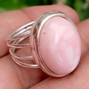 May include: A silver ring featuring a large, smooth, oval-shaped light pink stone set in a polished metal bezel. The band is constructed from four thin, parallel silver wires that converge at the base of the stone setting, creating a layered look.