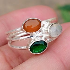 May include: A stackable silver ring set featuring three distinct bands, each topped with an oval-shaped gemstone. The stones include a smooth orange carnelian, a translucent white moonstone, and a faceted deep green emerald-coloured stone.