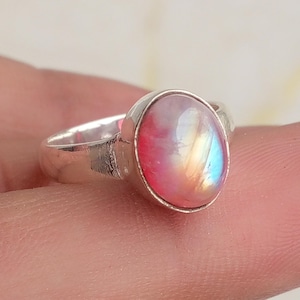 May include: A silver ring featuring a smooth, oval-shaped cabochon gemstone. The stone displays a translucent quality with iridescent flashes of pink, orange, and soft blue hues. The metal band is polished and has a simple, rounded design.