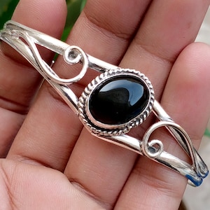Peut inclure: Un bracelet manchette en argent orné d'une pierre noire ovale centrale sertie dans une bordure en corde torsadée. La bande métallique présente des volutes décoratives de chaque côté de la pierre, créant un design ouvert au fini poli.