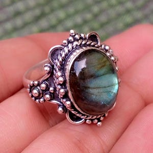 May include: A silver ring featuring an oval-shaped labradorite gemstone with blue and green iridescence. The metal setting is decorated with intricate twisted rope patterns and small spherical beads surrounding the central stone.