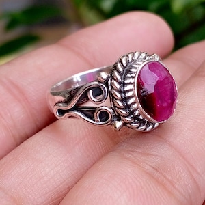 Puede incluir: Un anillo de plata con una piedra preciosa central de color rosa intenso, forma ovalada y tallada. La banda de metal presenta un borde intrincado similar a una cuerda trenzada alrededor de la piedra y patrones de volutas en los laterales.