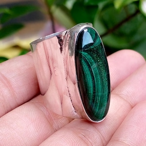 May include: A wide silver ring featuring a large, oval-shaped malachite gemstone. The stone displays deep green tones with characteristic swirling, banded patterns. The metal band is smooth, polished, and has a substantial, thick construction.